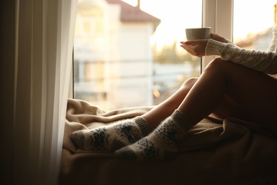 Photo of Woman in knitted socks holding cup of hot drink on plaid near window at home, closeup