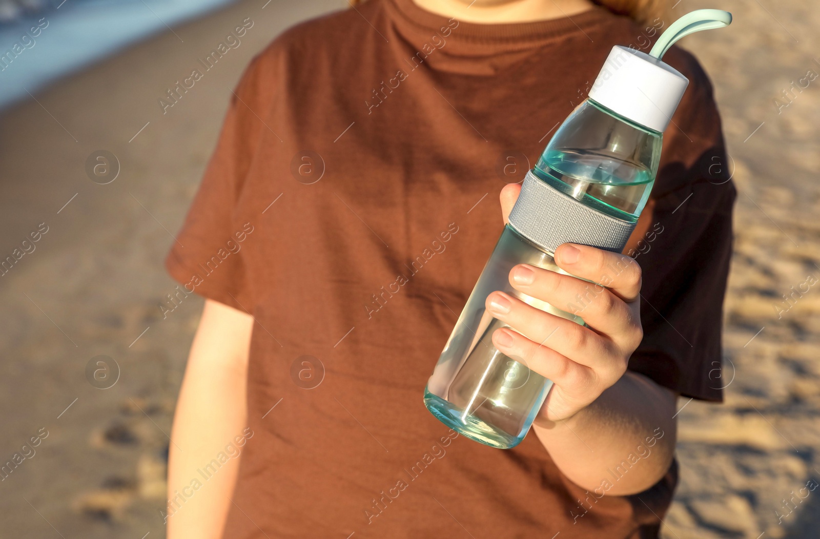 Photo of Woman holding glass bottle with water on sandy beach, closeup