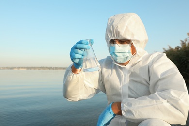 Scientist in chemical protective suit with conical flask taking sample from river for analysis