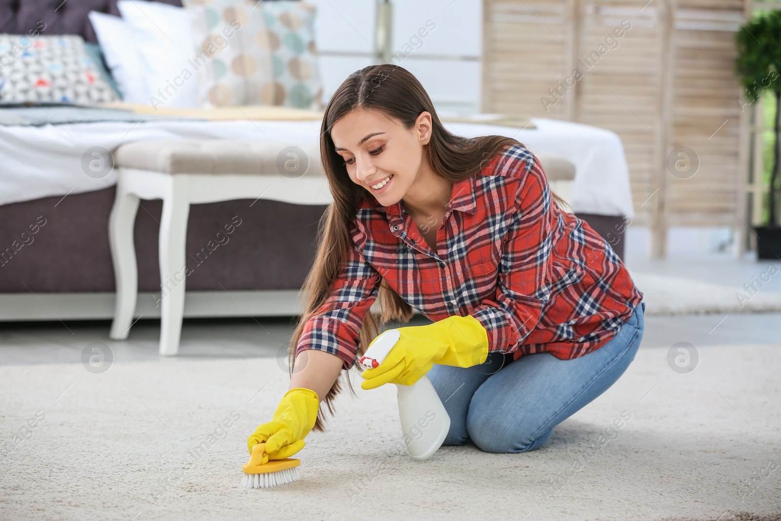 Photo of Young woman cleaning carpet with brush in bedroom