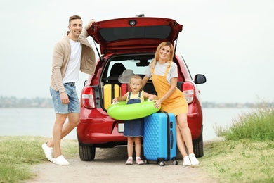 Happy family with suitcases and inflatable ring near car on riverside