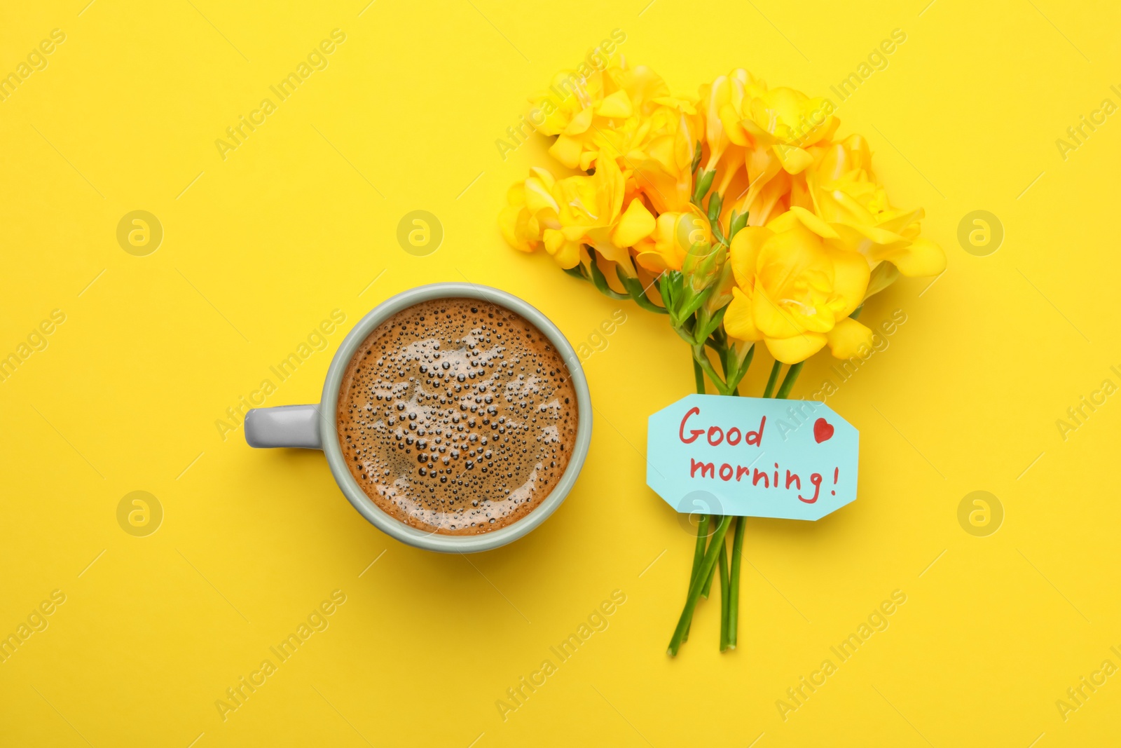 Photo of Cup of aromatic coffee, beautiful freesias and Good Morning note on yellow background, flat lay