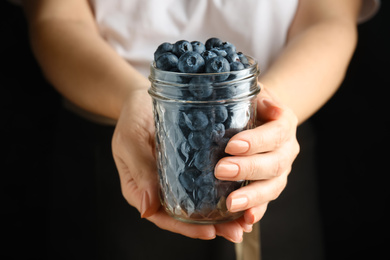 Woman holding jar with juicy fresh blueberries on black background, closeup