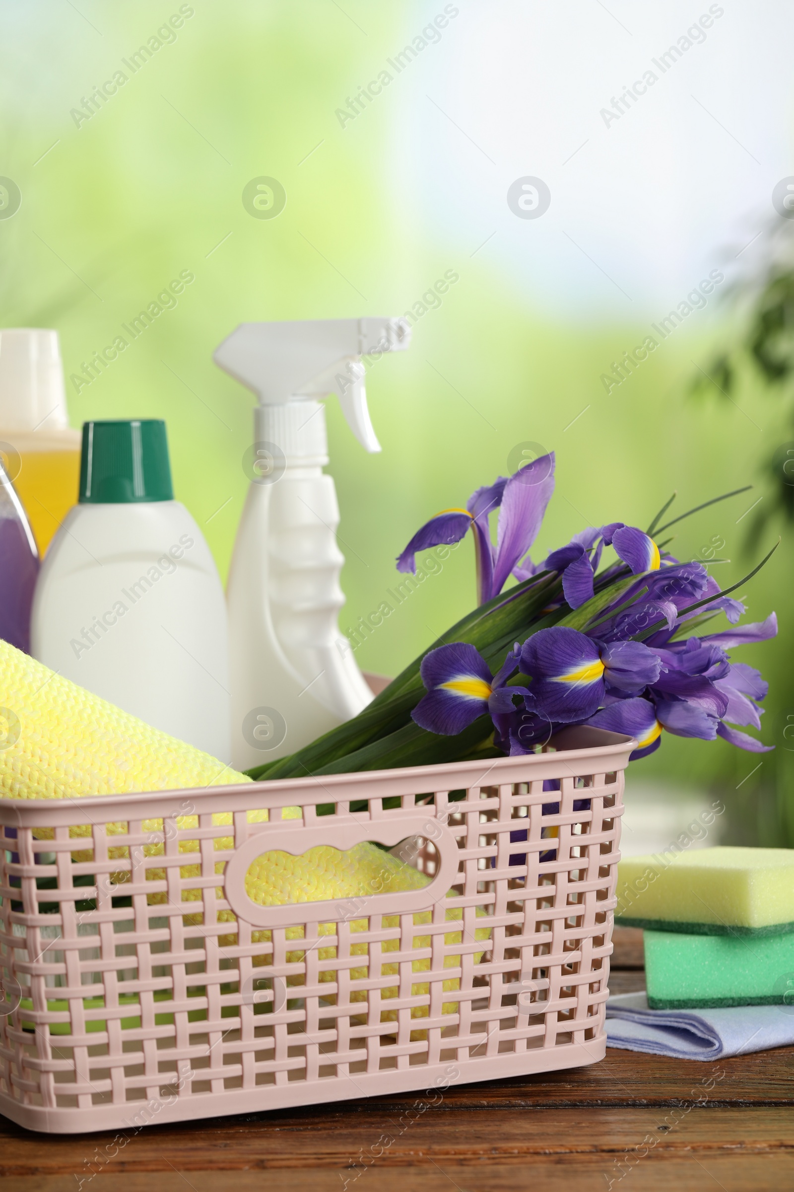 Photo of Spring cleaning. Plastic basket with detergents, supplies and beautiful flowers on wooden table outdoors