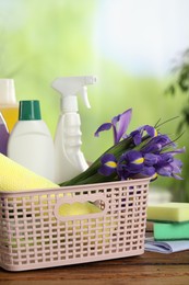 Photo of Spring cleaning. Plastic basket with detergents, supplies and beautiful flowers on wooden table outdoors