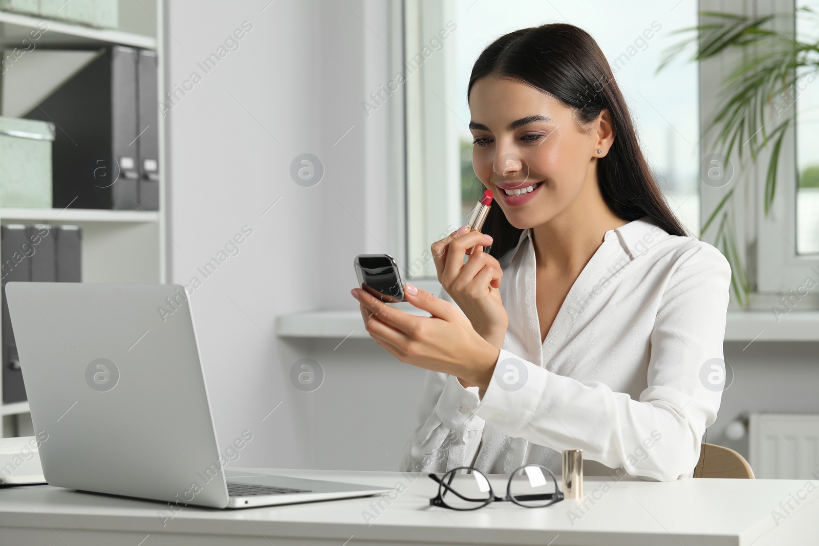 Photo of Young woman using cosmetic pocket mirror while applying makeup in office