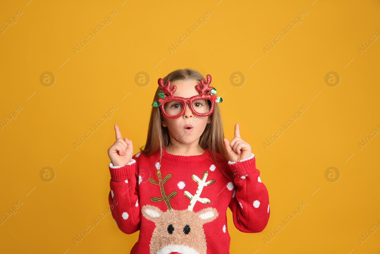 Photo of Surprised little girl in Christmas sweater and party glasses pointing on yellow background