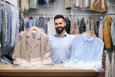 Photo of Dry-cleaning service. Happy worker holding hangers with clothes in plastic bags at counter indoors