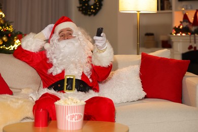 Photo of Merry Christmas. Santa Claus changing TV channels with remote control on sofa at home