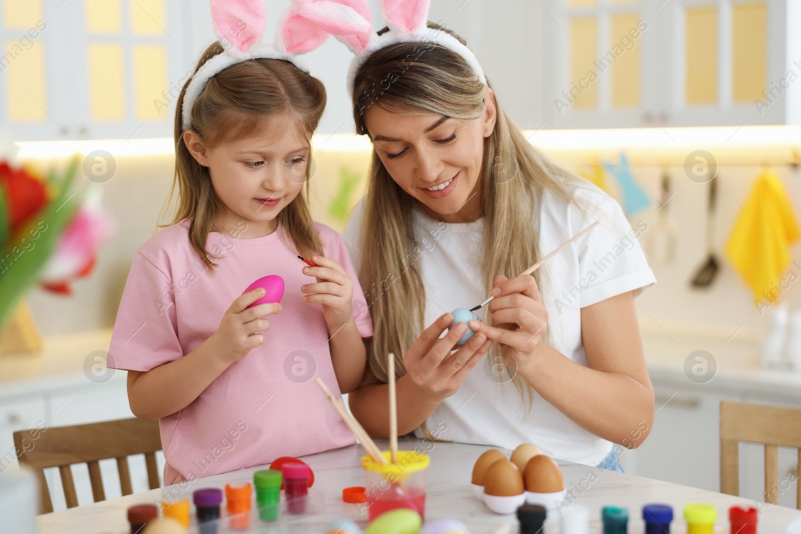 Photo of Easter celebration. Mother with her cute daughter painting eggs at white marble table in kitchen