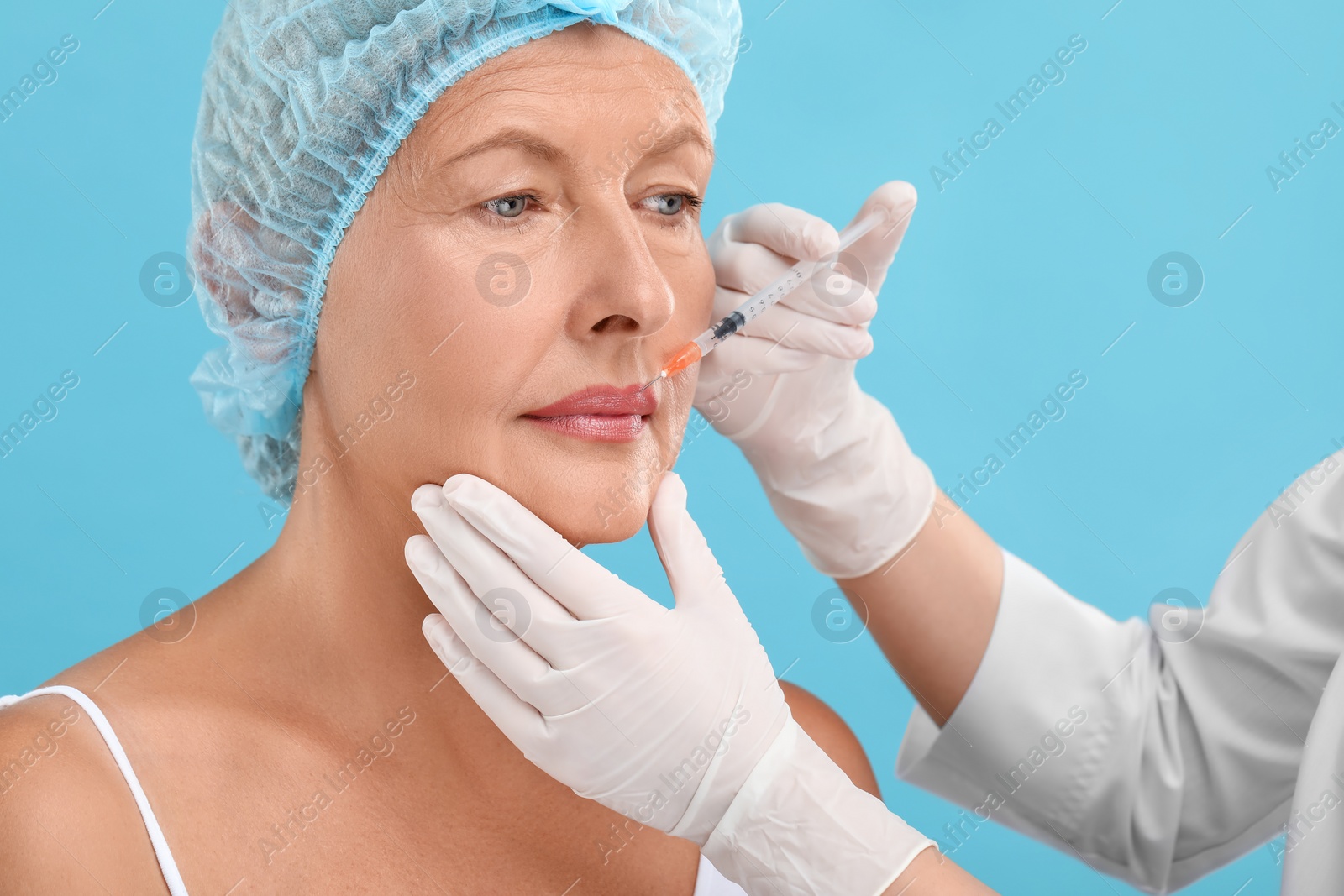 Photo of Doctor giving lips injection to senior woman on light blue background. Cosmetic surgery