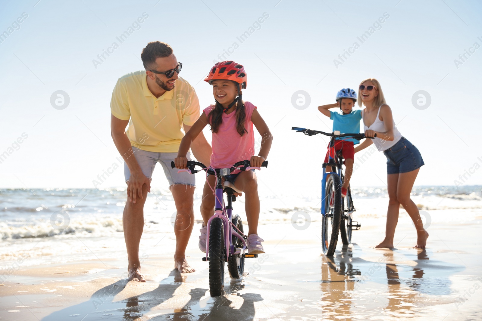 Photo of Happy parents teaching children to ride bicycles on sandy beach near sea