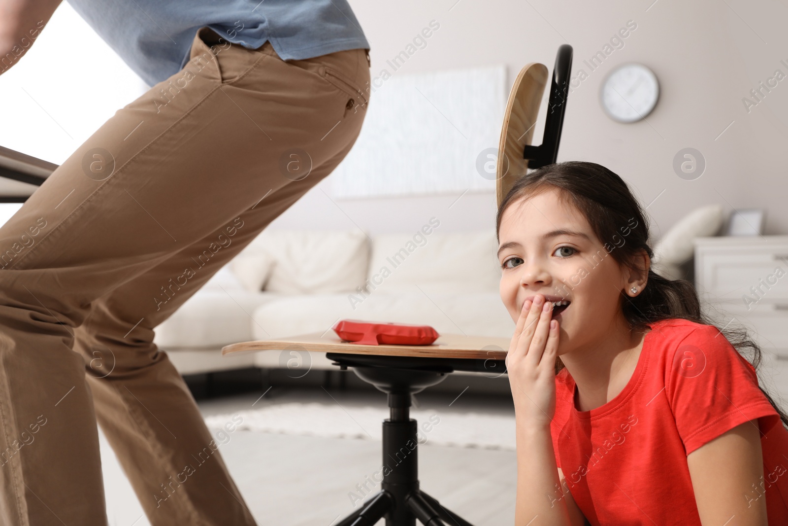 Photo of Cute little girl putting whoopee cushion on father's chair while he sitting down at home, closeup