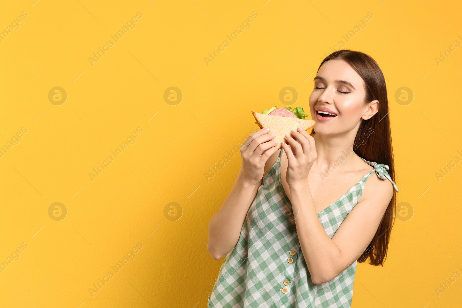 Photo of Young woman eating tasty sandwich on yellow background. Space for text