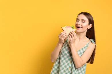 Young woman eating tasty sandwich on yellow background. Space for text