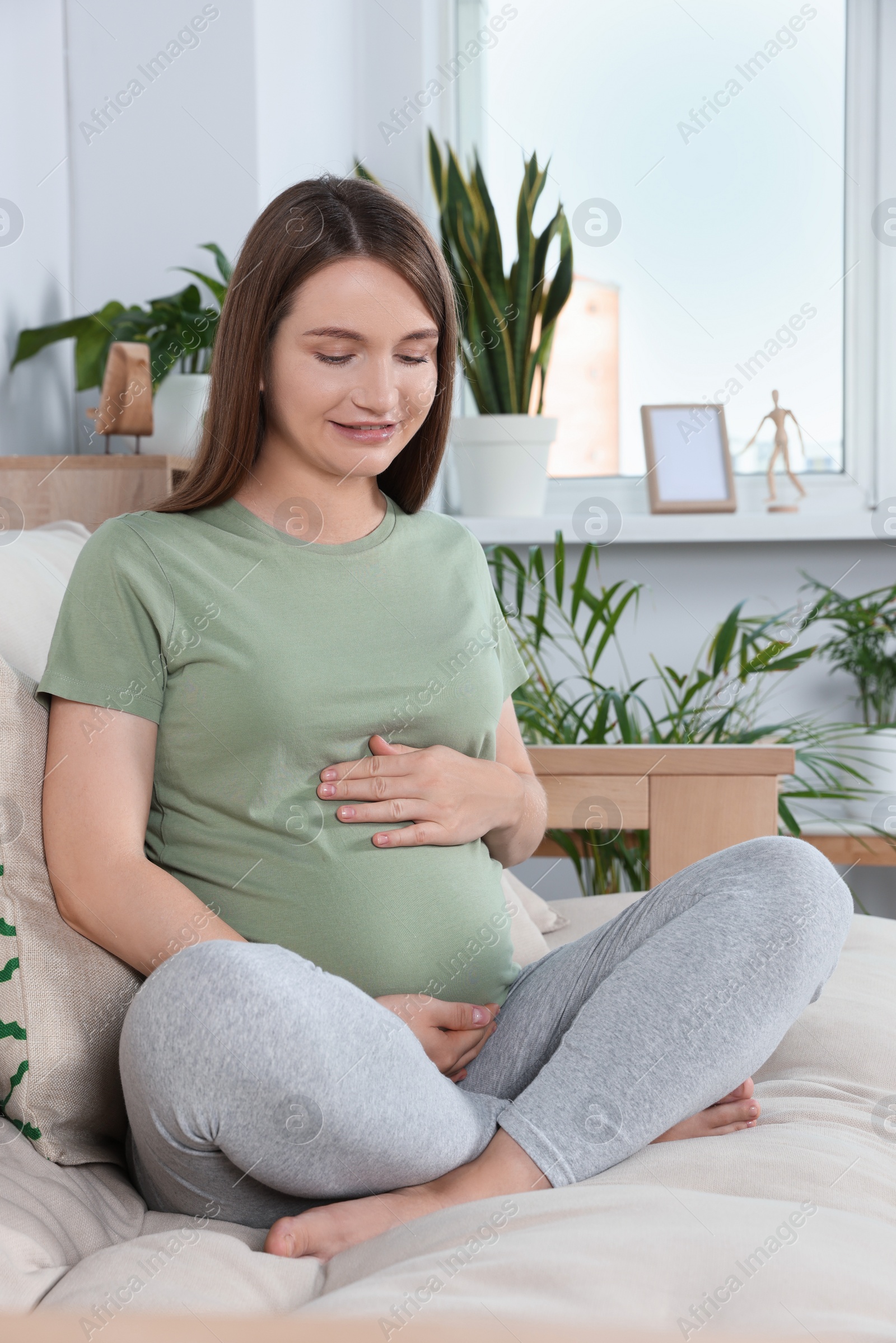 Photo of Happy pregnant woman sitting on sofa and touching her belly in living room