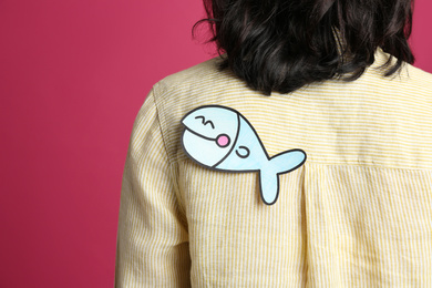 Photo of Woman with paper fish on back against pink background, closeup. April fool's day