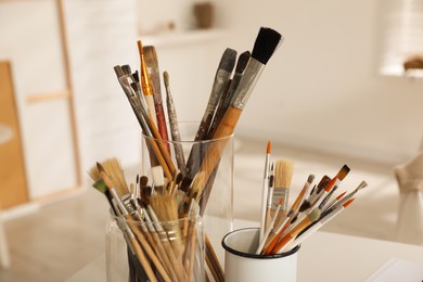 Photo of Holders with different paintbrushes on white table in studio, closeup. Artist's workplace