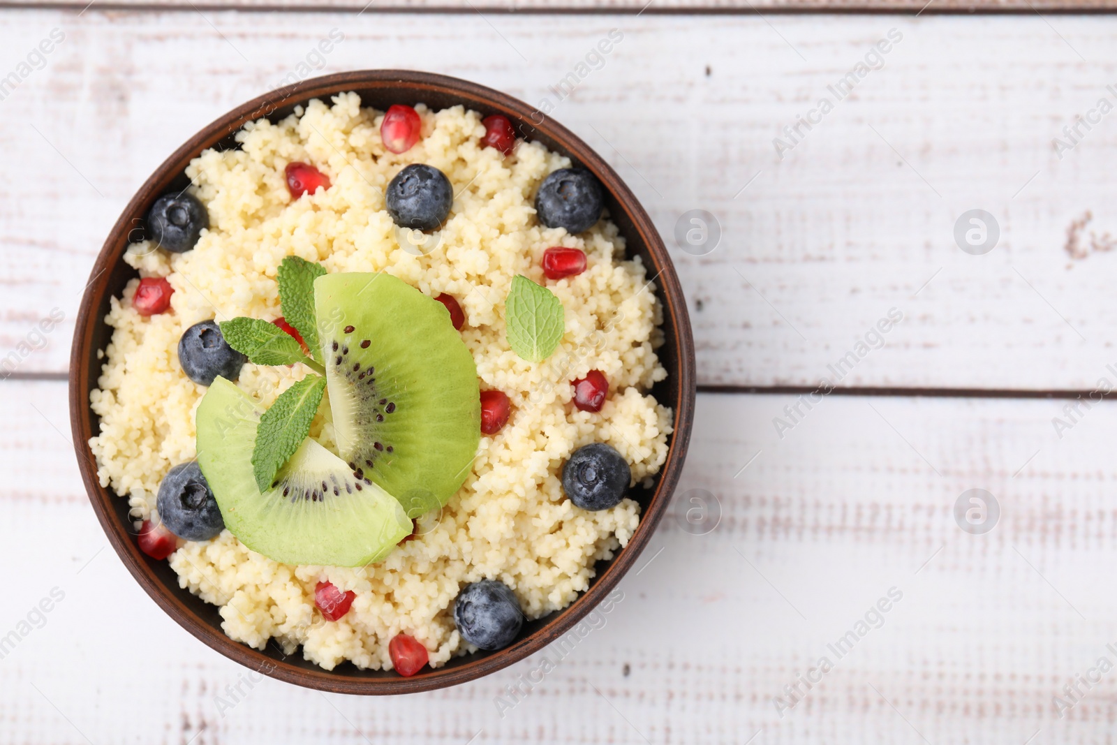 Photo of Bowl of tasty couscous with kiwi, blueberries and pomegranate on white wooden table, top view. Space for text