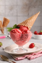 Photo of Delicious scoops of strawberry ice cream with mint and wafer cone in glass dessert bowl served on grey table