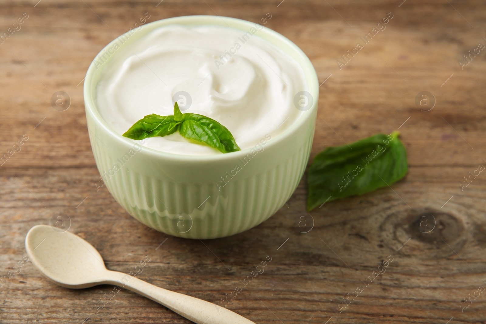 Photo of Bowl of fresh sour cream with basil and spoon on wooden table