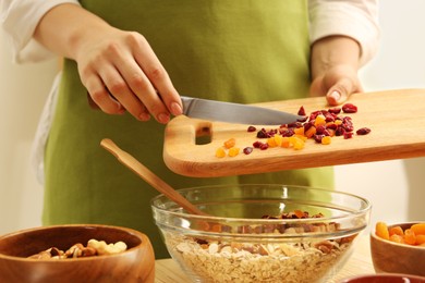 Making granola. Woman adding dried apricots and cherries into bowl with oat flakes at table in kitchen, closeup