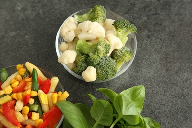 Bowls of different frozen vegetables and fresh basil on grey table, above view
