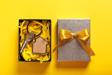 Key with trinket in shape of house and gift box on yellow background, flat lay. Housewarming party