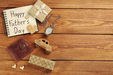 Photo of Notebook with phrase HAPPY FATHER'S DAY and male accessories on wooden table, flat lay. Space for text
