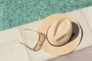 Stylish hat and sunglasses near outdoor swimming pool on sunny day, above view. Beach accessories