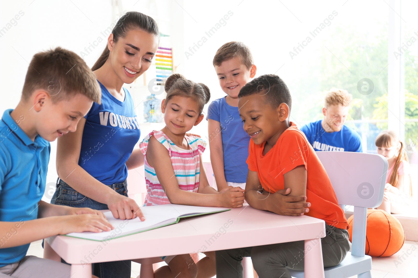 Photo of Young volunteer reading book with children at table indoors