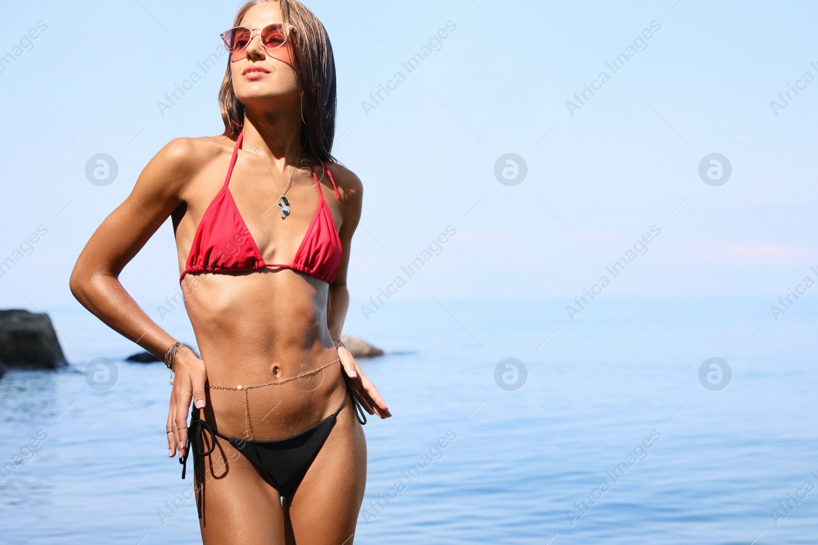 Photo of Beautiful young woman in stylish bikini and sunglasses on seashore, space for text