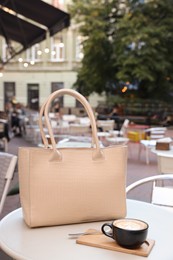 Photo of Stylish bag and cup of coffee on white table in outdoor cafe