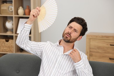 Photo of Bearded man waving white hand fan to cool himself on sofa at home