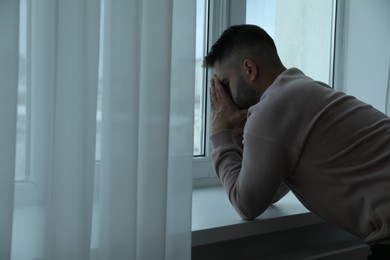 Photo of Sad man near window at home. Space for text