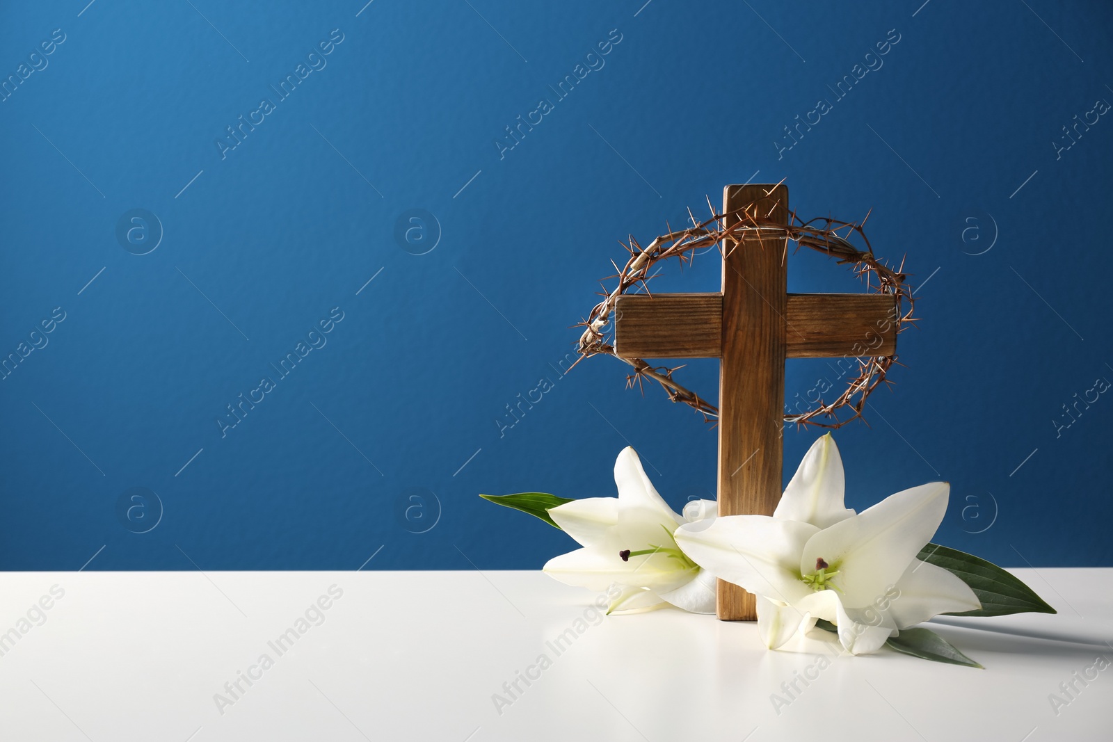 Photo of Wooden cross, crown of thorns and blossom lilies on table against color background, space for text