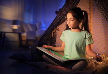 Photo of Little girl reading fairy tale near play tent in room