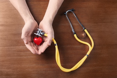 Photo of Man holding model of heart and stethoscope on wooden table. Heart attack concept