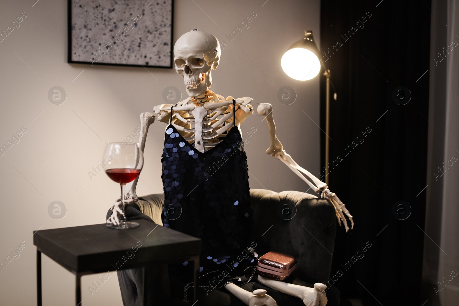 Photo of Skeleton in dress with wine sitting at table indoors