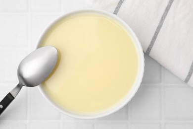 Photo of Bowl with condensed milk and spoon on white tiled table, flat lay
