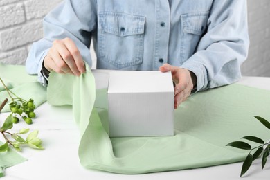 Photo of Furoshiki technique. Woman wrapping gift in green fabric at white table, closeup