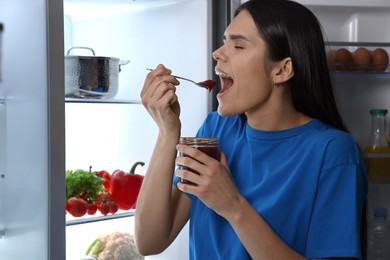 Young woman eating sweet jam near modern refrigerator at night