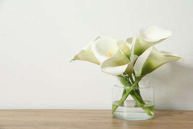 Photo of Beautiful calla lily flowers in glass vase on wooden table near white wall. Space for text