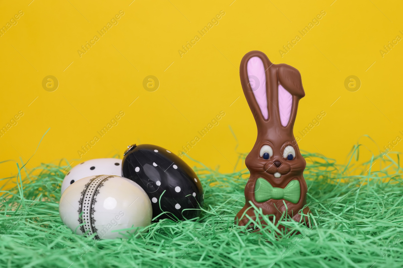 Photo of Easter celebration. Cute chocolate bunny and painted eggs on grass against yellow background, space for text