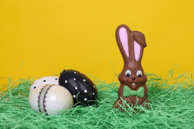 Easter celebration. Cute chocolate bunny and painted eggs on grass against yellow background, space for text