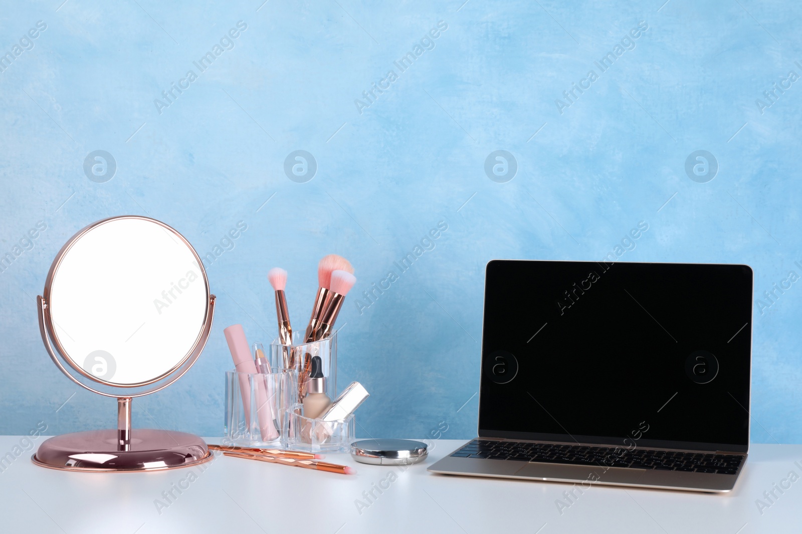 Photo of Modern laptop, mirror and makeup products on table near light blue wall. Space for design