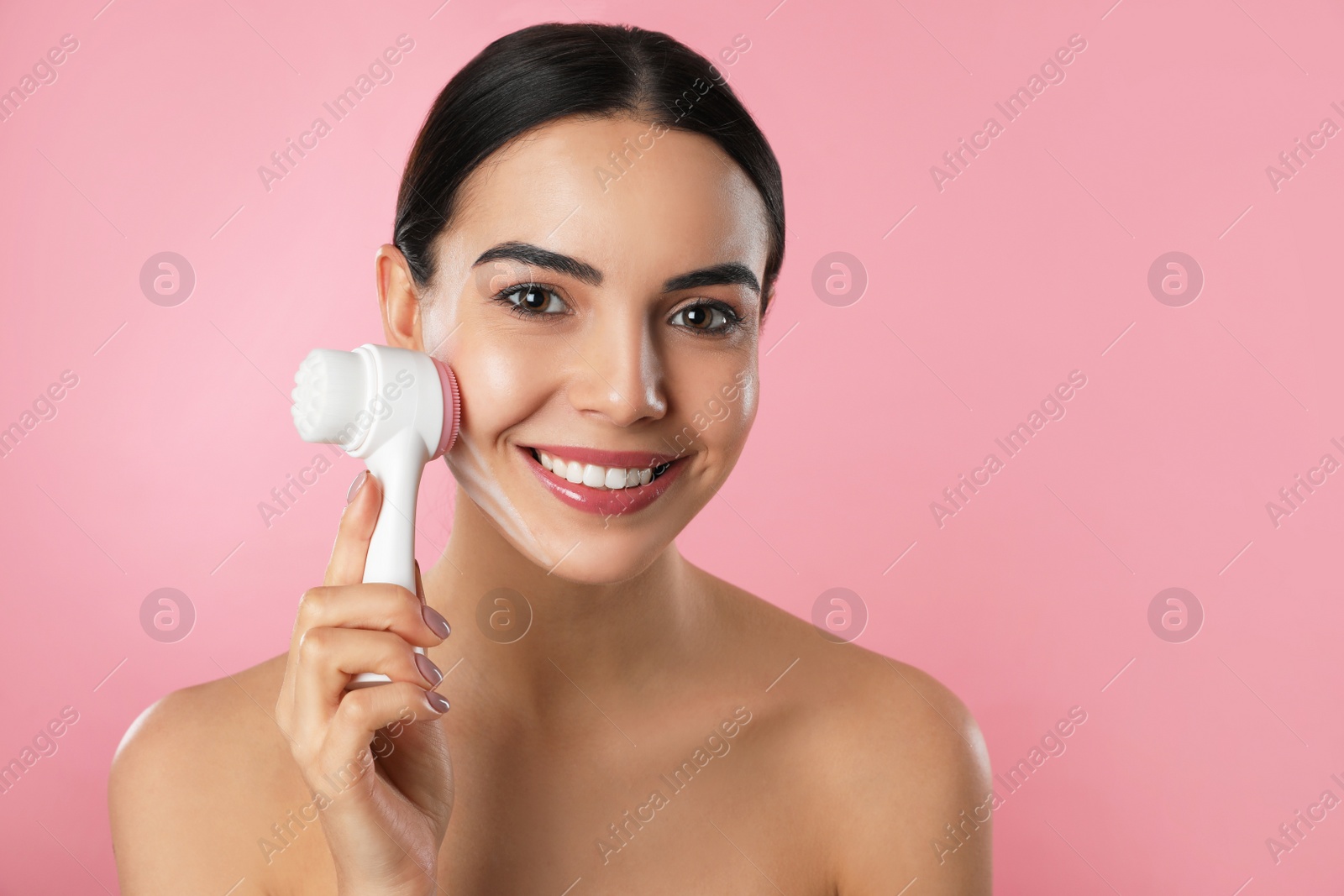 Photo of Young woman using facial cleansing brush on pink background. Washing accessory