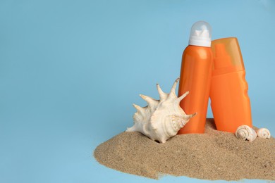 Photo of Sand with bottles of sunscreens and seashells against light blue background, space for text. Sun protection