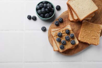 Photo of Delicious toasts with peanut butter and blueberries on white tiled table, top view. Space for text