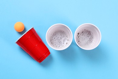 Plastic cups and ball for beer pong on light blue background, flat lay